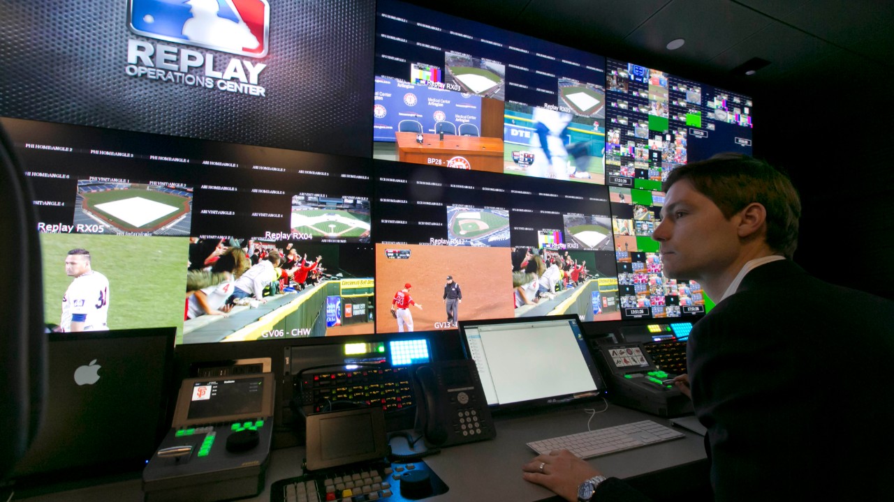 Man in a black suit watching multiple MLB broadcast screens