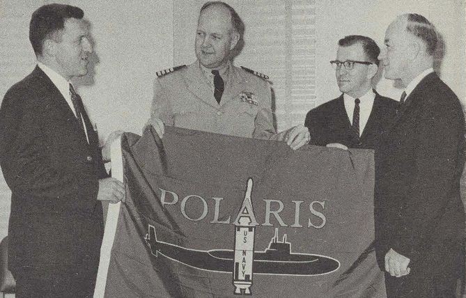 Four men in suits hold a flag labeled “Polaris” and illustrated with a submarine and a rocket ship