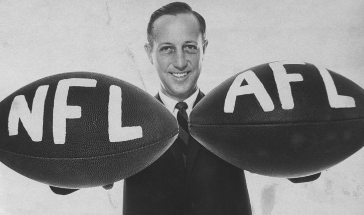 Clean cut man in a suit smiles while holding two footballs labelled “NFL” and “AFL”