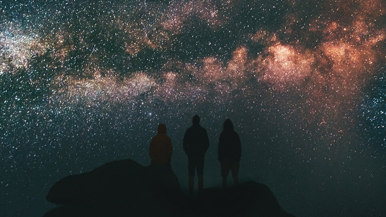 A group of three people standing on a hill and looking at the night sky.